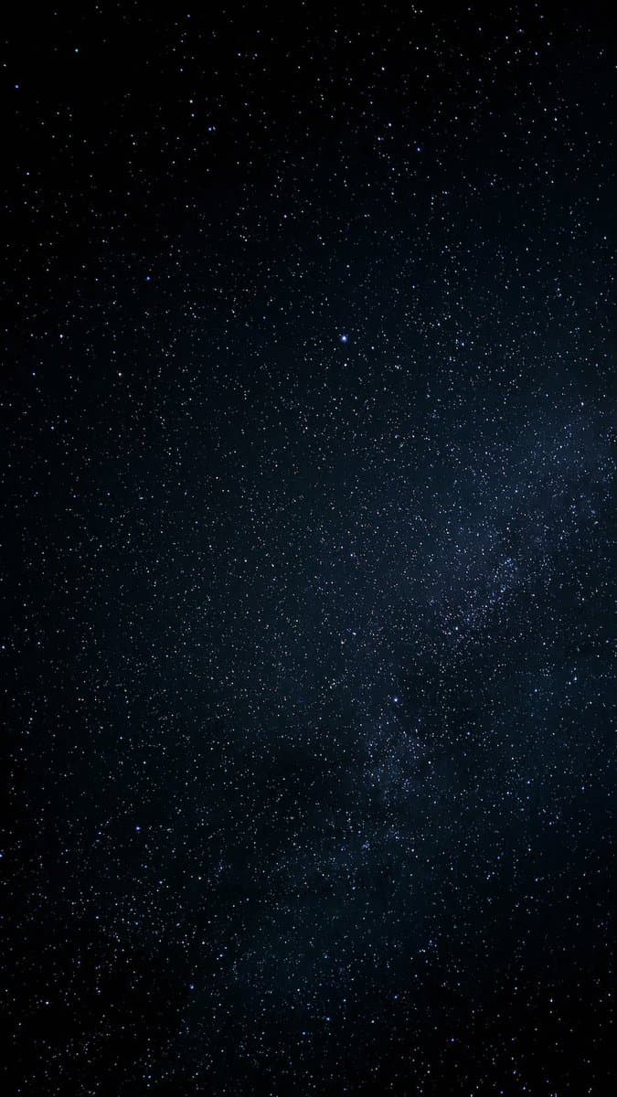 A photograph of a starry night sky, showing a dense field of stars against a dark background. A faint, milky band of light, likely the Milky Way, is visible stretching diagonally across the image.