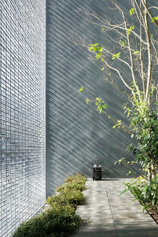 A photograph of a modern architectural exterior featuring a textured glass-block wall on the left and a smooth, dark gray wall on the right. A small garden path with low hedges runs between the walls, leading to a black lantern, with a leafy tree and shrubbery on the right side.