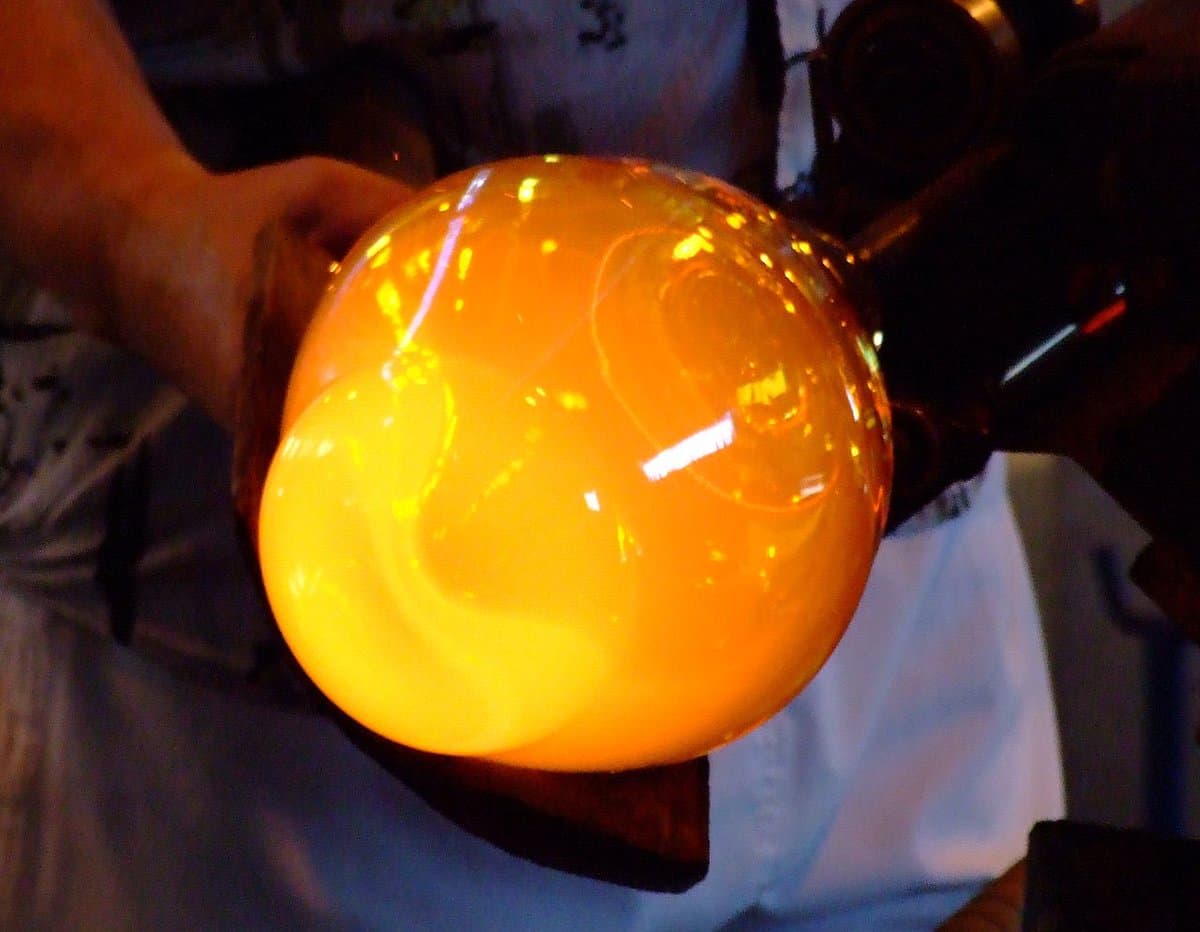 A close-up photograph of a person holding a large, glowing orange glass sphere, likely a piece of glass art or a lamp, in a dimly lit indoor setting.