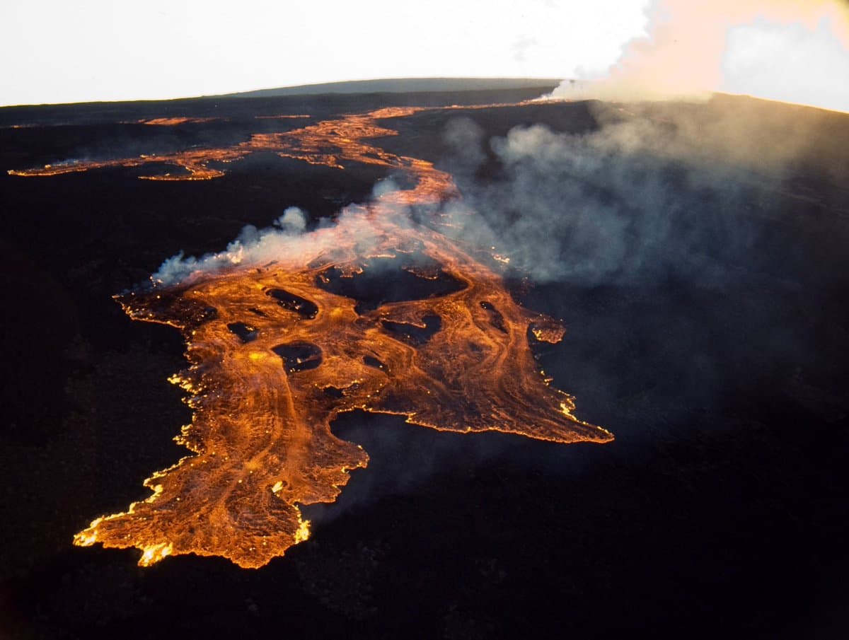 An aerial photograph showing a volcanic eruption with bright orange lava flowing across a dark landscape, accompanied by rising plumes of white and grey smoke.