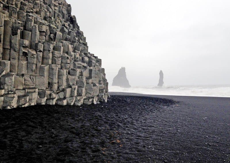 A coastal landscape featuring a black sand beach in the foreground and a large, textured cliff face made of hexagonal basalt columns on the left. In the misty background, two sea stacks rise from the ocean, with waves crashing against the shore.