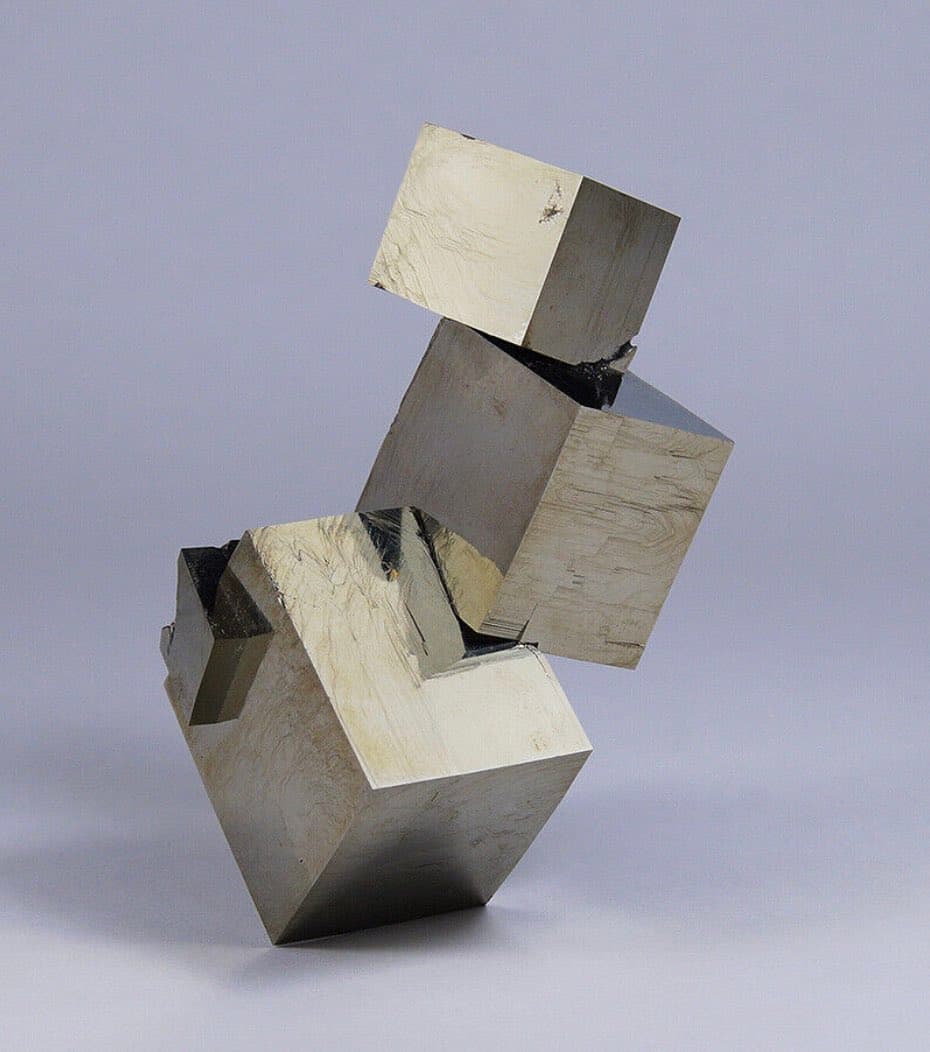 A studio photograph of an abstract geometric sculpture made of stacked, polished metal blocks with a brushed finish, set against a plain light gray background.