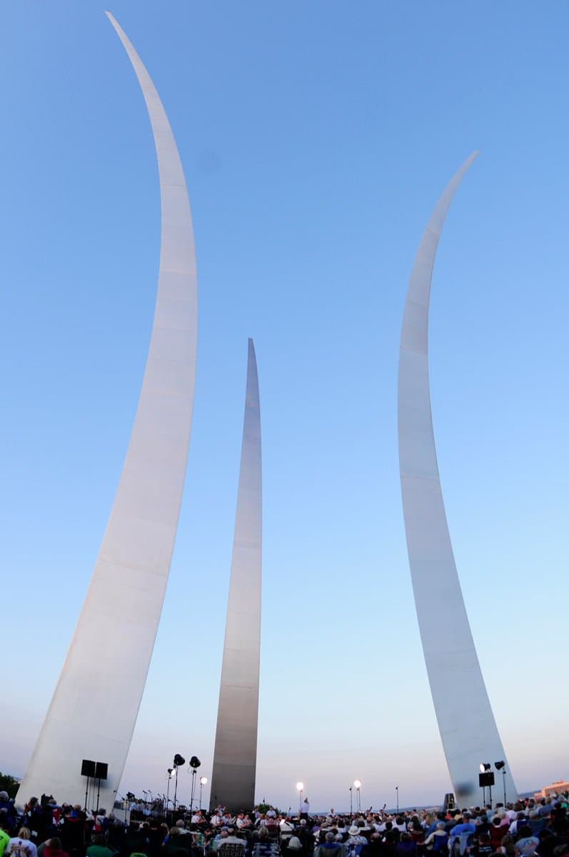A low-angle photograph of the Gateway Arch in St. Louis, Missouri, showing its three tall, curved stainless steel legs against a clear blue sky. A large crowd of people is gathered at the base of the monument, with stage lighting and speakers visible, suggesting a public event or concert.