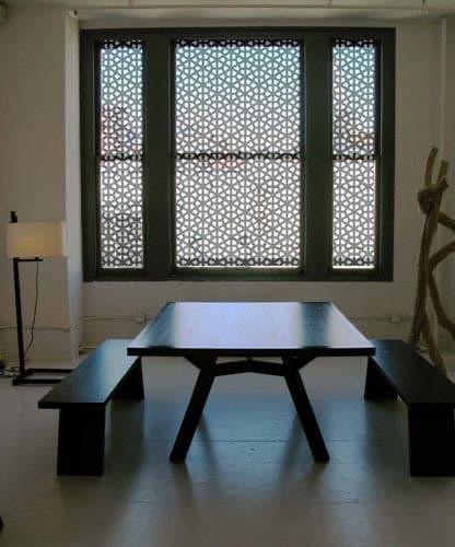 A minimalist dining room featuring a long wooden table with matching benches, positioned in front of a large window covered with decorative geometric lattice screens. A floor lamp stands to the left, and a sculptural wooden piece is visible on the right.