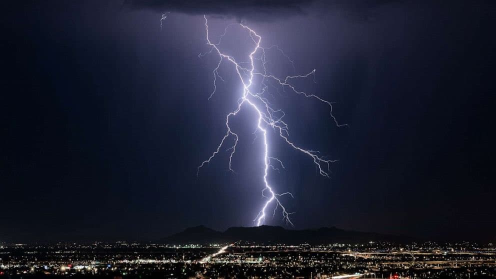A dramatic nighttime photograph captures a massive lightning bolt striking the ground over a city skyline. The bright, branching lightning illuminates the dark clouds above, while the city lights below provide a contrasting foreground.