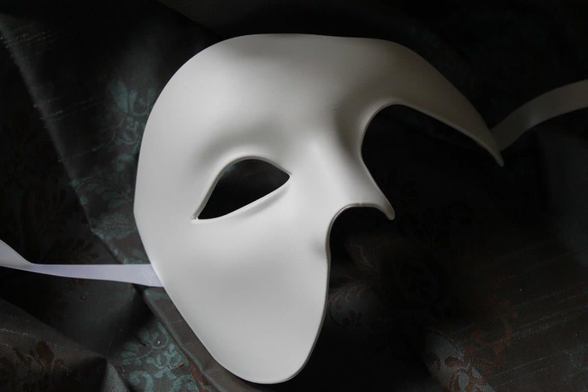 A white, blank masquerade mask with a pointed nose and eye holes rests on a dark, patterned fabric. The mask has a smooth, matte finish and is attached to a white ribbon.