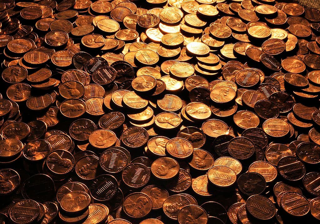 A close-up photograph of a large pile of U.S. one-cent coins (pennies) stacked and scattered together, illuminated by warm, golden light. The image shows the obverse (Lincoln's profile) and reverse (Lincoln Memorial) sides of the coins.