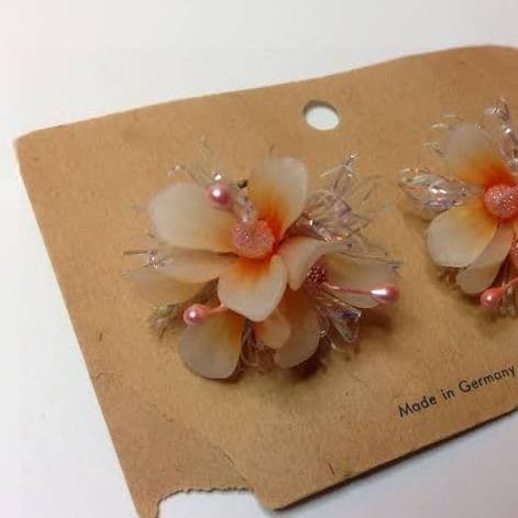 A pair of decorative, flower-shaped earrings with pearl and crystal accents displayed on a brown cardboard backing card. The card has a small hole at the top and text printed on the bottom right corner.