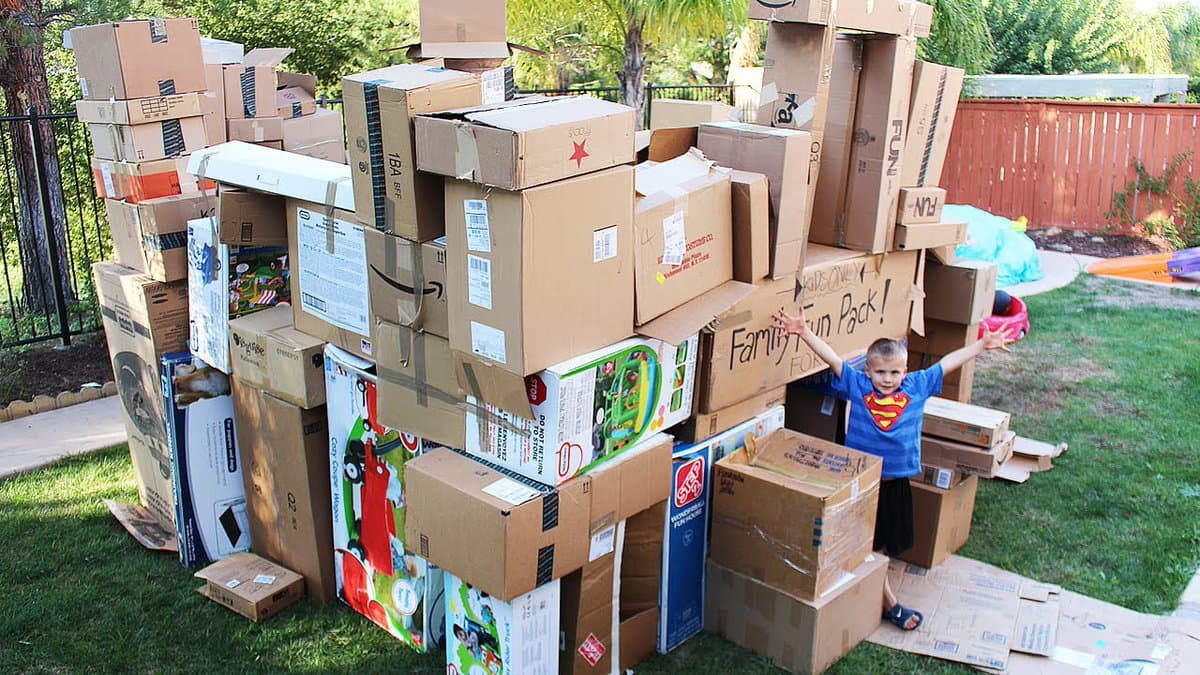 A young boy in a Superman t-shirt stands with arms raised next to a large, precarious stack of cardboard boxes in a backyard. The boxes, many bearing Amazon logos and shipping labels, are piled high on the grass, with one box labeled "Family Fun Pack!".