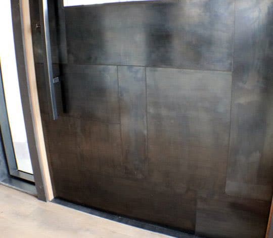 A close-up view of a dark, textured metal door or wall panel with a vertical handle on the left side, next to a window frame.