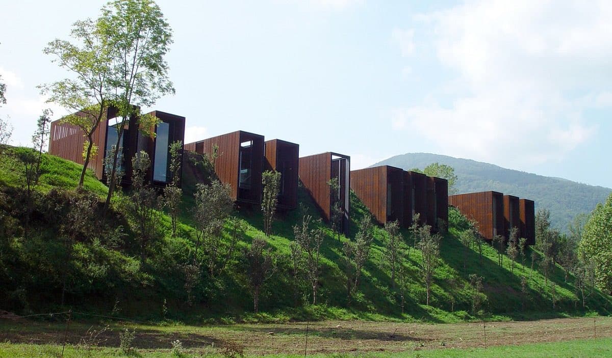 A row of modern, rust-colored rectangular cabins is built into a grassy hillside, with young trees planted in front of them and a mountain visible in the background under a partly cloudy sky.