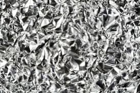 A black-and-white close-up photograph of crumpled, reflective material, likely aluminum foil or a similar metallic surface. The image shows a chaotic texture of sharp creases and folds with varying levels of brightness and shadow.