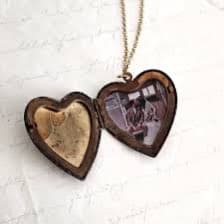 A heart-shaped locket necklace with a gold chain, shown open to reveal a small photograph inside one half and a gold interior in the other, resting on a white surface with faint cursive writing.