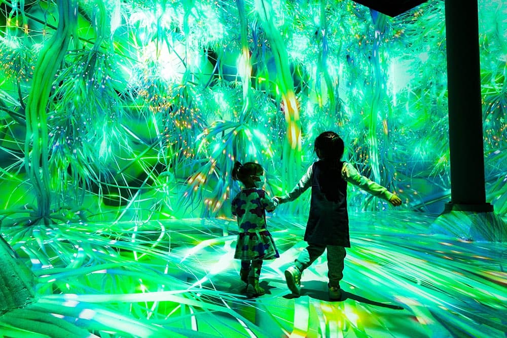 Two children walk through an immersive digital art installation featuring vibrant, glowing green projections of grass and foliage on the walls and floor.