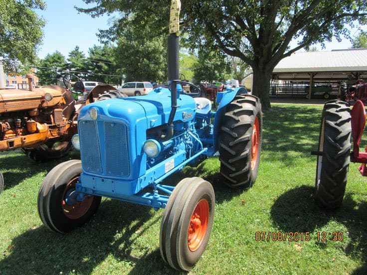 A vintage blue Farmall tractor with orange wheels is parked on a grassy lawn at an outdoor event, with other antique farm equipment visible in the background. The image includes a digital timestamp in the bottom right corner.