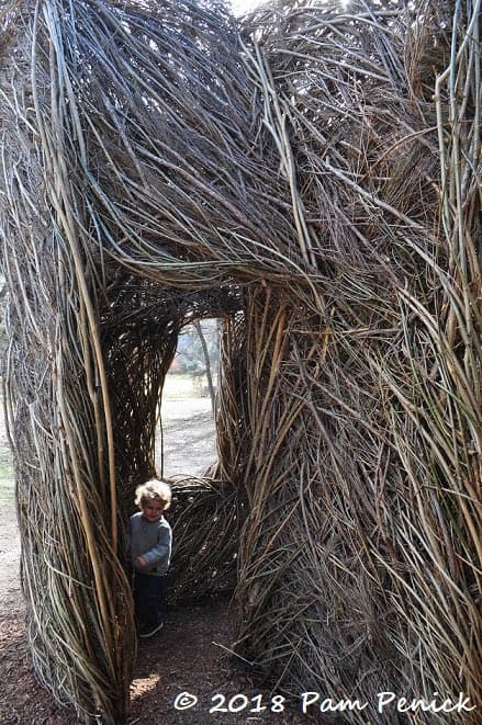 A young child stands inside a large, open-walled structure made of woven branches and twigs, resembling a rustic hut or sculpture.