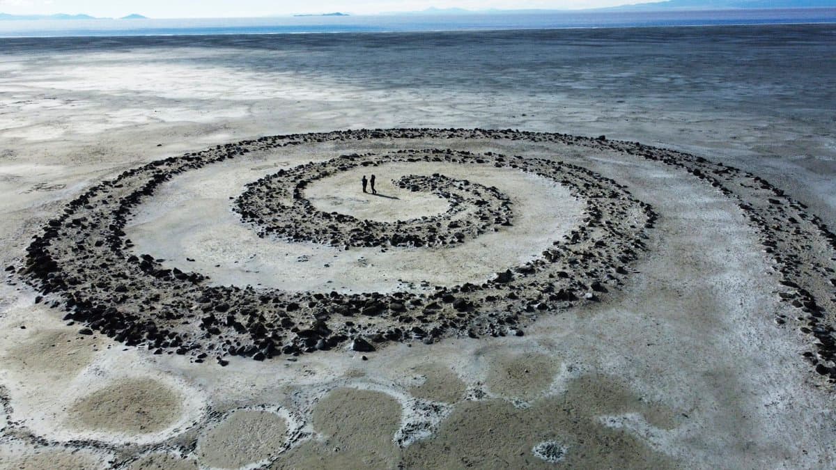 An aerial photograph of the Spiral, a large stone art installation on a vast, flat salt flat landscape. Two small figures stand near the center of the spiral, providing a sense of the structure's immense scale against the expansive, arid terrain.