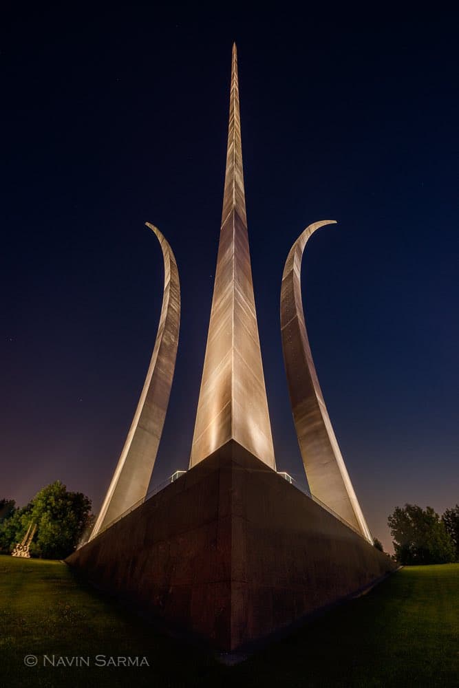 A low-angle night photograph of the Air Force Memorial, featuring a tall central spire flanked by two curved wings illuminated against a dark sky. The monument stands on a large stone base surrounded by grass and trees.