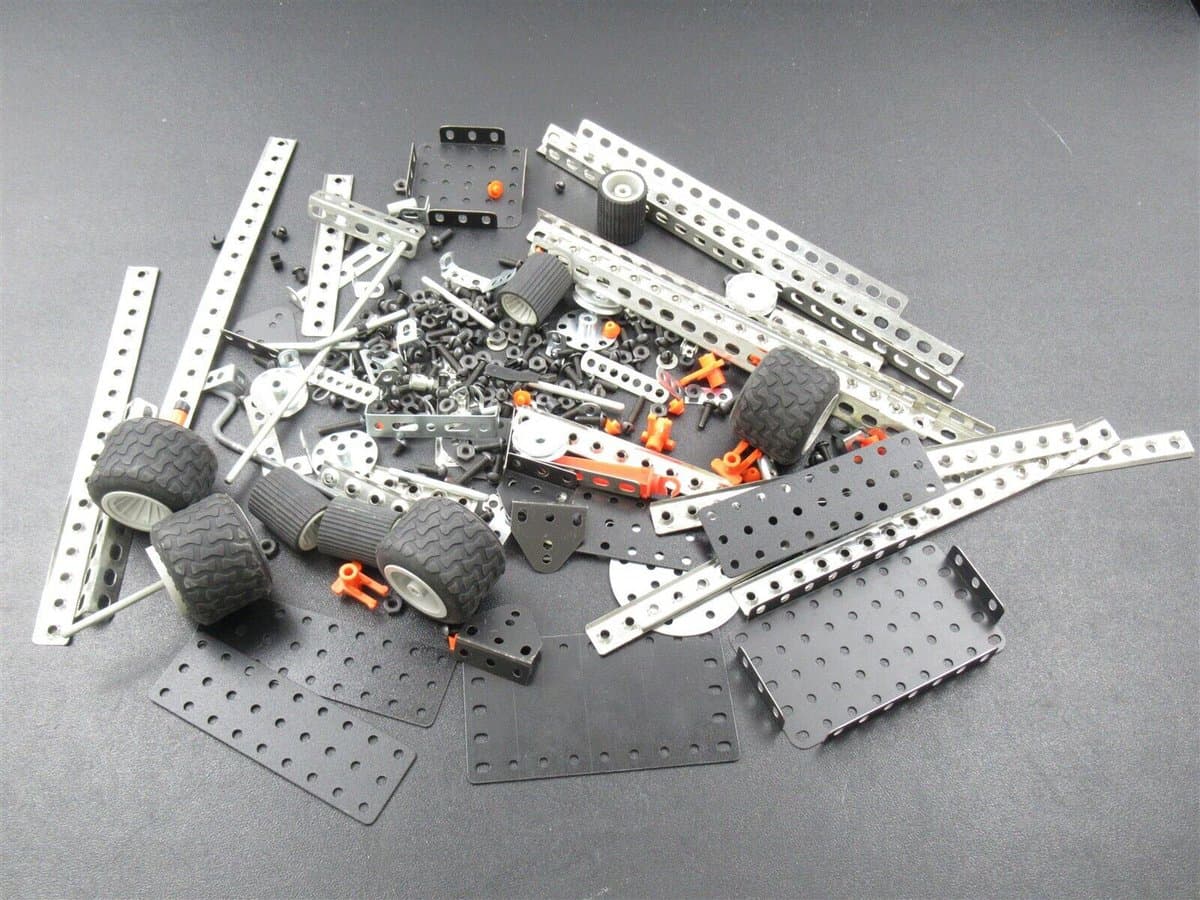 A collection of disassembled robot parts, including metal beams, black plates, wheels, gears, and fasteners, laid out on a dark surface.