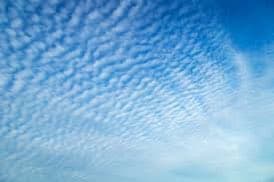 A photograph of a bright blue sky filled with a pattern of small, white, rippled clouds known as altocumulus. The clouds create a textured, wave-like appearance across the upper portion of the image, fading into a lighter, hazier sky near the bottom.