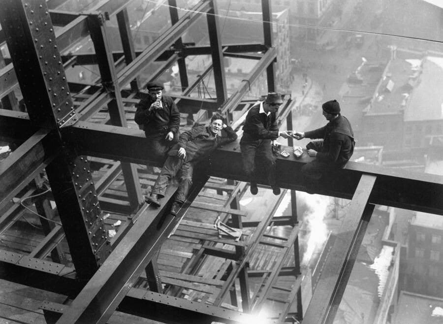 A black-and-white historical photograph showing four construction workers sitting and lying on a steel beam high above a city, with one man smoking and others sharing a meal.