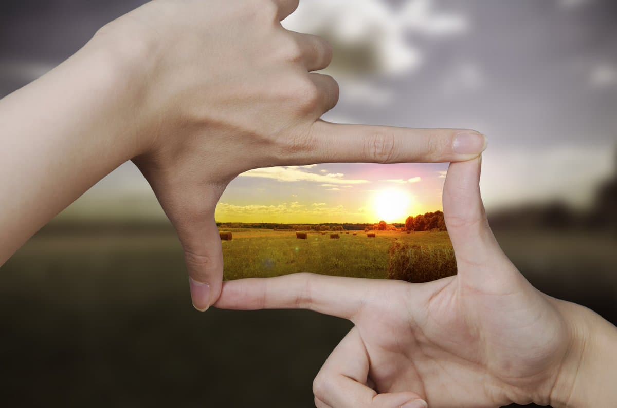 A pair of hands forms a rectangular frame with the thumb and index finger, revealing a small image of a sunset over a field with hay bales. The background is a blurred, overcast landscape.