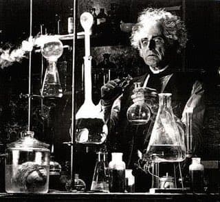 A black-and-white photograph of an older man with curly hair standing in a laboratory, holding a glass flask and surrounded by various scientific glassware and equipment.