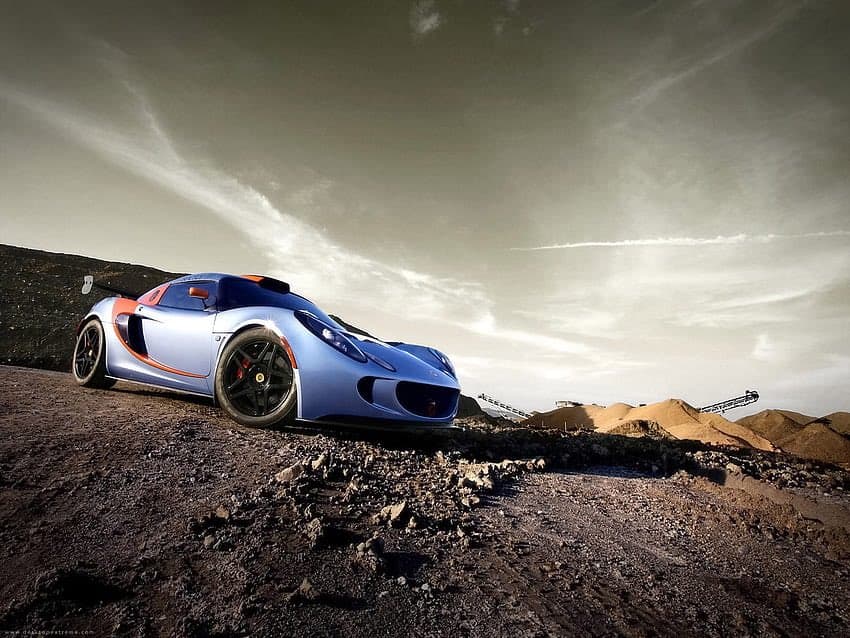 A low-angle photograph of a blue and orange McLaren P1 GTR supercar parked on a rocky, gravel-covered hillside under a dramatic, cloudy sky. In the background, there are piles of earth and industrial conveyor belts, suggesting a quarry or construction site.