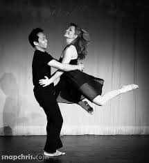 A black-and-white photograph of a man and a woman performing a dance lift on a stage. The man is supporting the woman, who is extended horizontally in the air with one leg stretched out. A watermark reading 'snapchris.com' is visible in the bottom left corner.
