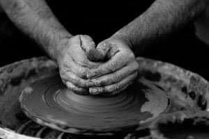 A black-and-white photograph showing a pair of hands shaping wet clay on a spinning pottery wheel. The image captures the tactile process of pottery making, with the hands firmly pressing into the rotating material.