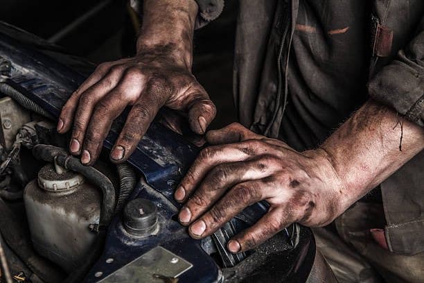 A close-up photograph showing a pair of hands covered in black grease and grime working on the engine of a vehicle.