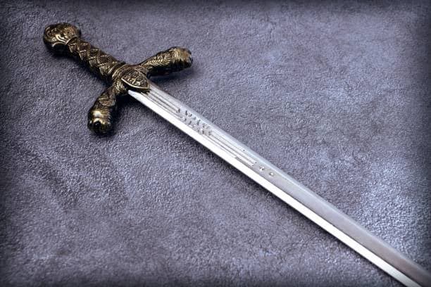 A close-up photograph of a decorative sword with an ornate, gold-colored hilt and a silver blade resting on a textured gray surface. The blade features engraved text near the hilt. The image has a dark, vignetted border.