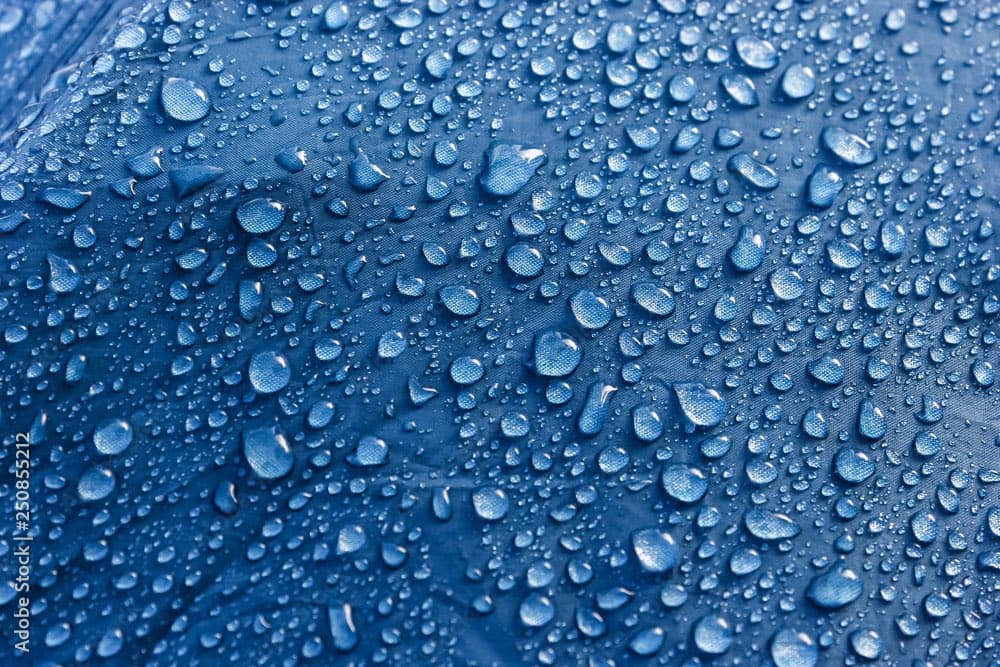 A close-up photograph of a blue fabric surface covered in numerous water droplets of varying sizes.