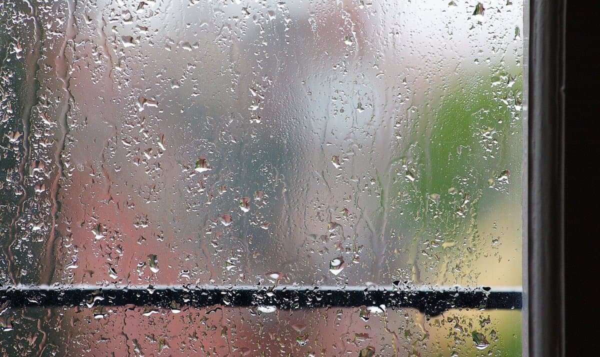 A close-up photograph of a window pane covered in rain droplets, with a blurred view of a red building and green foliage visible in the background.