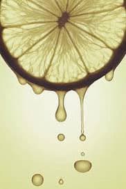 A close-up photograph of a sliced lemon with juice dripping from the bottom edge, forming droplets against a pale yellow background.