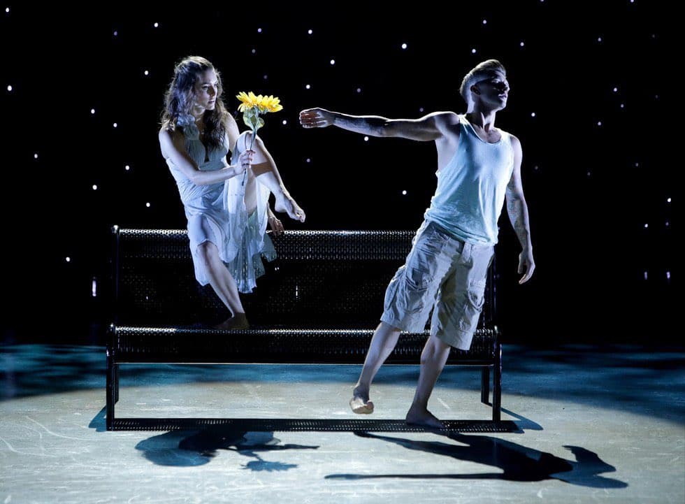 A photograph of two dancers performing on a stage with a black background featuring small white lights. A woman in a white dress sits on a bench holding a yellow flower, while a man in a white tank top and shorts stands beside her with his arm extended.