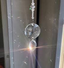 A close-up photograph of a crystal prism hanging from a beaded chain, refracting sunlight into a bright starburst pattern against a window background.