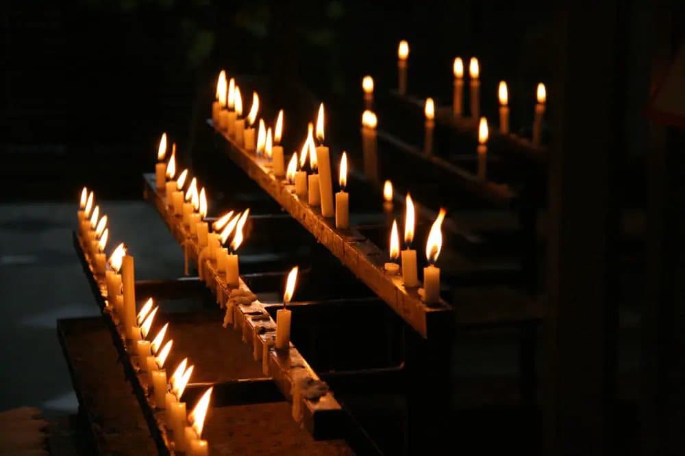 A photograph of numerous lit white candles arranged in rows on wooden racks in a dark setting, likely a church or memorial site. The warm candlelight illuminates the immediate area while the background remains in shadow.