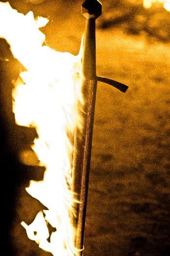 A sword is shown standing upright with a large fire burning on its left side, set against a dark, textured background. The image has a warm, golden-brown color tone.
