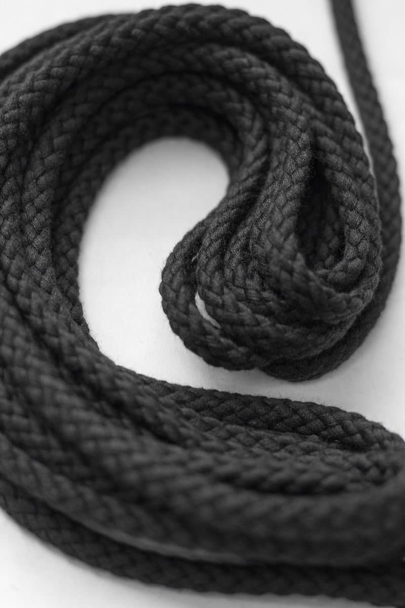 A close-up, black-and-white photograph of a thick, braided black rope coiled in a loose loop on a white surface. The image focuses on the texture of the rope's weave and the shadows within its curves.