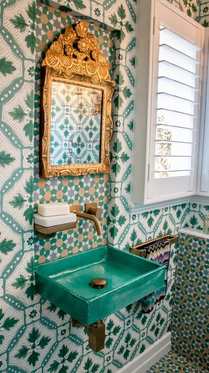 A bathroom interior featuring a green ceramic sink, a gold-framed mirror, and a window with white shutters, all set against walls covered in green and white patterned wallpaper. The room has a vintage, ornate aesthetic with a brass faucet and towel rack.