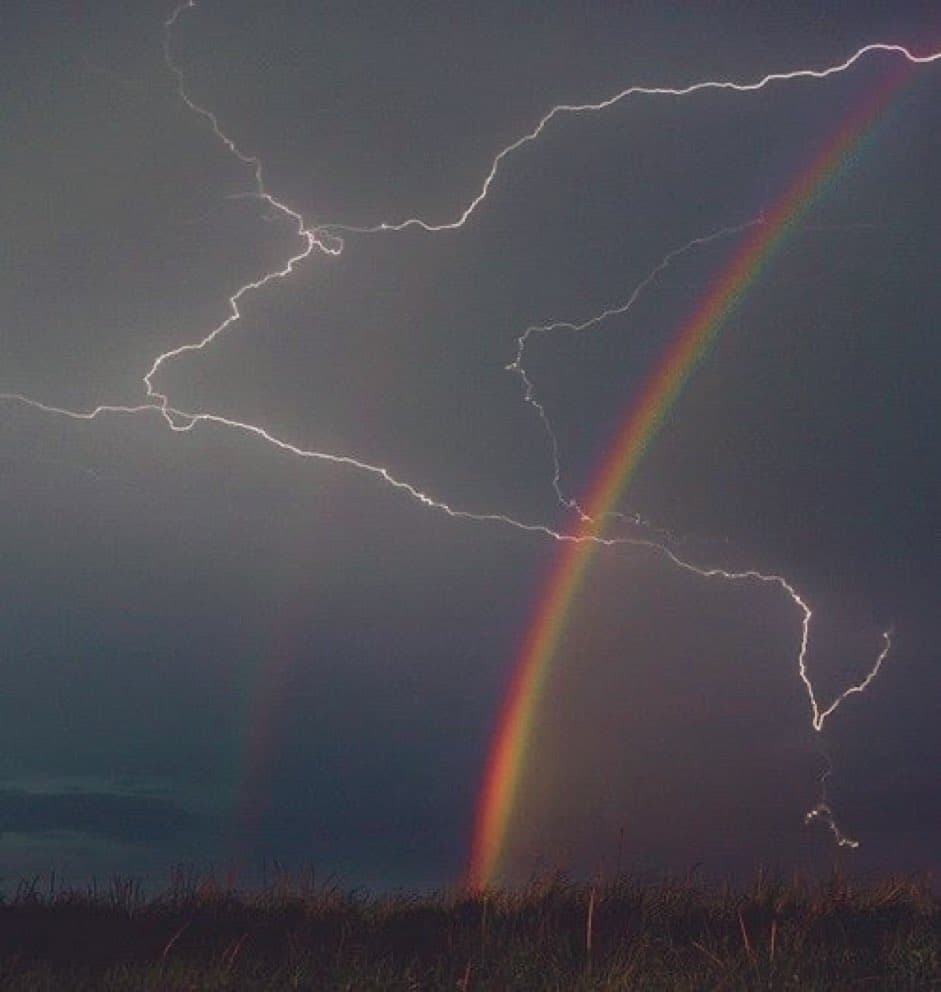 A dramatic photograph of a dark stormy sky featuring bright, branching lightning bolts striking across the frame. A vivid rainbow arcs vertically through the center, intersecting the lightning, with a silhouette of a grassy field along the bottom edge.