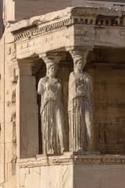 A close-up photograph of two ancient Greek caryatid statues serving as architectural columns on a stone building facade. The figures are carved in a standing pose with draped clothing, supporting an entablature above them.