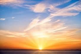 A scenic photograph of a sunset over a body of water, with the sun low on the horizon casting bright rays through a sky filled with wispy clouds. The sky transitions from deep blue at the top to warm orange and yellow near the horizon. The water reflects the colors of the sky in the foreground.