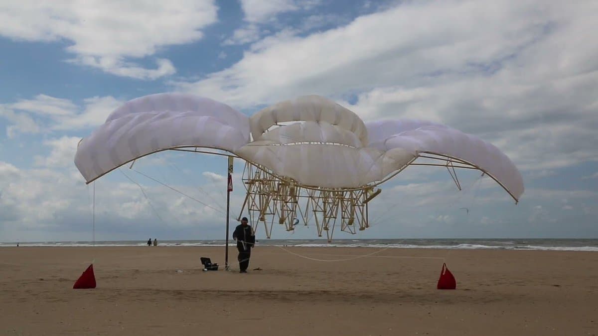 A large, white, multi-winged experimental aircraft or hang glider is being set up on a sandy beach under a partly cloudy sky. A person in dark clothing stands near the structure, which is supported by a complex framework and tethered to the ground with red markers.