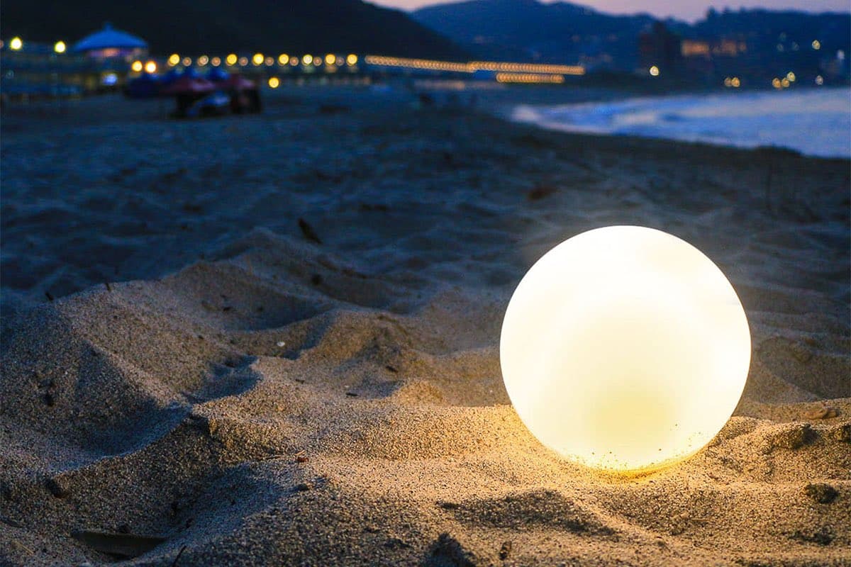 A glowing, spherical light sits on a sandy beach at dusk, casting a warm glow on the sand. In the blurred background, there is a shoreline with distant lights and a body of water.