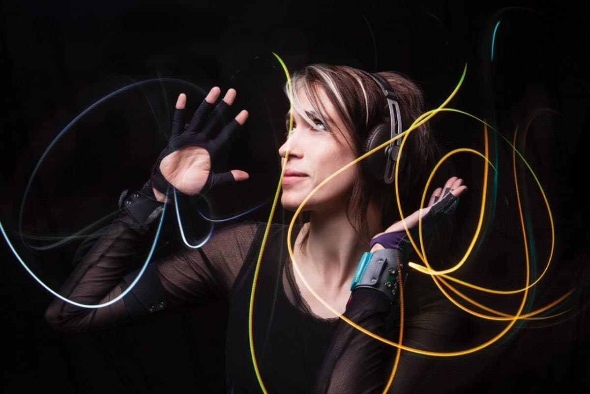 A woman wearing headphones and fingerless gloves poses against a black background, surrounded by swirling trails of colored light created by long-exposure photography. She is looking to the side with her hands raised, as if conducting or interacting with the light patterns.