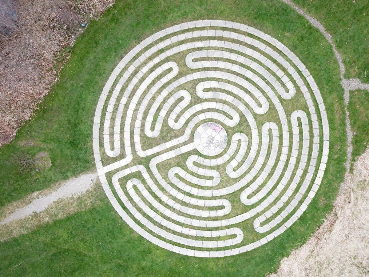 An aerial view of a circular stone labyrinth set into a green lawn, with a single entrance path leading to the center. The surrounding area includes patches of dry leaves and a gravel path.