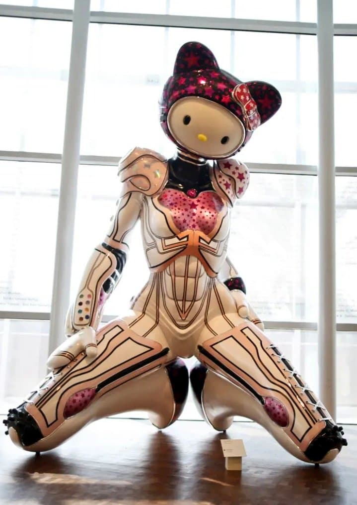 A photograph of a large, glossy sculpture of the character Hello Kitty wearing a futuristic, white and pink armored suit. The figure is posed in a wide, crouching stance on a wooden floor in front of a large window.