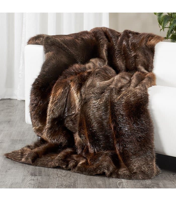 A thick, brown faux-fur throw blanket is draped over the arm and seat of a white sofa, with the excess fabric pooling on the gray wood-look floor. The setting includes a white curtain in the background and a green plant to the right.